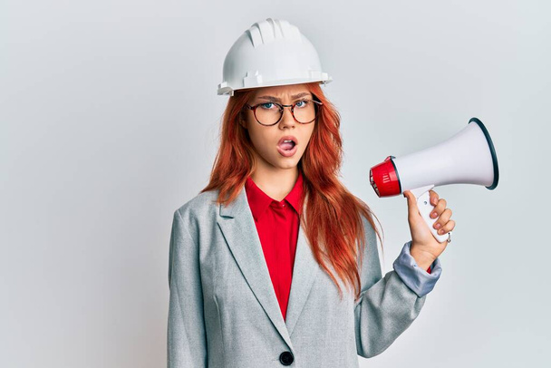 Молодая рыжая женщина в архитектурной каске и мегафоне в шоке, выглядит скептически и саркастично, удивлена с открытым ртом  - Фото, изображение