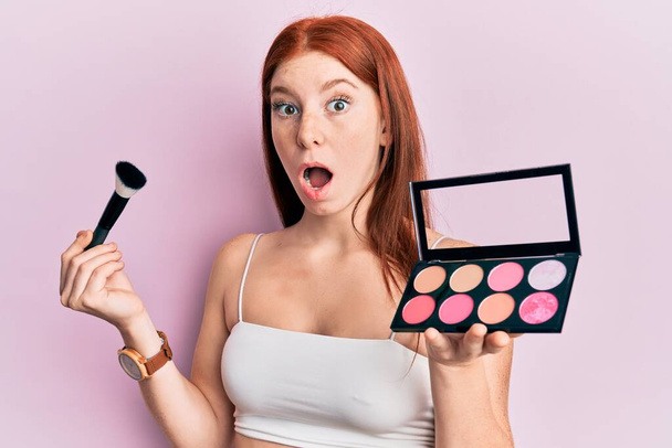 Young red head girl holding makeup brush and blush palette afraid and shocked with surprise and amazed expression, fear and excited face.  - Photo, Image