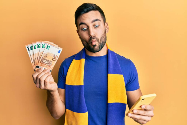 Молодий прихильник іспаномовного футболу за допомогою смартфона, який тримає банкноти в 50 євро, роблячи рибу обличчям з ротом і примхливими очима, божевільною і комічною
.  - Фото, зображення