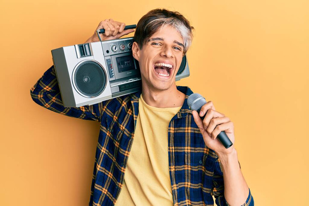 Jonge Spaanse man zingt lied met behulp van microfoon en boombox glimlachen en hard lachen, want grappige gekke grap.  - Foto, afbeelding