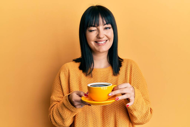 Junge hispanische Frau mit Kaffee zwinkernd in die Kamera blickend mit sexy Ausdruck, fröhlichem und glücklichem Gesicht.  - Foto, Bild