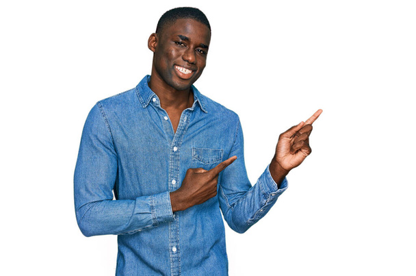 Fiatal afro-amerikai férfi alkalmi ruhában mosolyog és a kamerába néz, két kézzel és ujjal oldalra mutatva..  - Fotó, kép