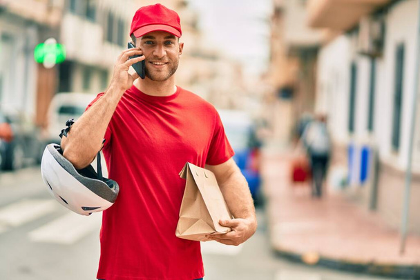 Jeune livreur caucasien parlant sur le smartphone et tenant sac en papier de livraison à la ville. - Photo, image