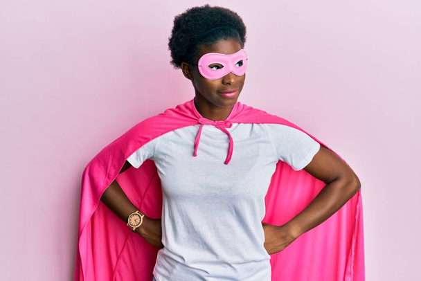 Молода афроамериканська дівчина в костюмі супергероя і медична маска посміхаються дивлячись в сторону і дивлячись геть думки.  - Фото, зображення