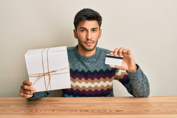 Fiatal jóképű férfi kezében ajándék és hitelkártya szkeptikus és ideges, homlokráncolás ideges, mert a probléma. negatív személy.  - Fotó, kép