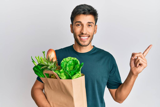 Jeune homme beau tenant sac en papier avec pain et épicerie souriant heureux pointant avec la main et le doigt sur le côté  - Photo, image