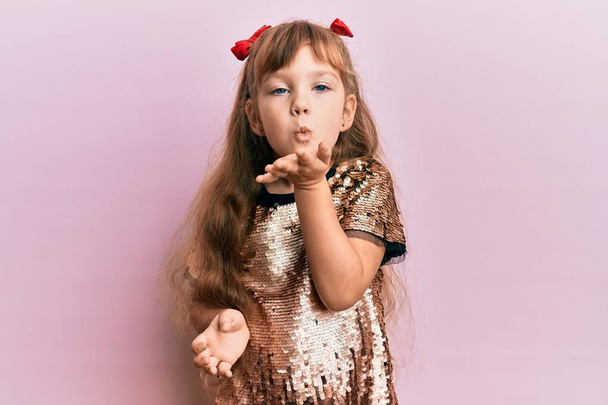 Petite fille caucasienne enfant portant robe de paillettes festives regardant la caméra souffler un baiser avec la main sur l'air étant belle et sexy. expression d'amour.  - Photo, image
