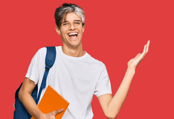 Young hispanic man wearing student backpack and holding books celebrating victory with happy smile and winner expression with raised hands  - Photo, Image