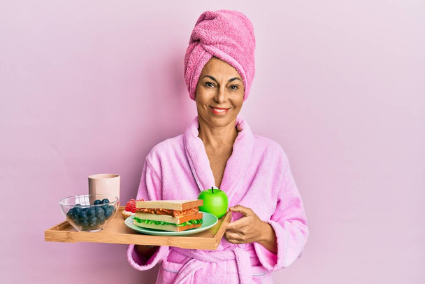 Mujer hispana de mediana edad con albornoz sosteniendo bandeja de desayuno con aspecto positivo y feliz de pie y sonriendo con una sonrisa confiada mostrando los dientes  - Foto, imagen