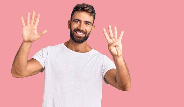 Joven hombre hispano vistiendo camiseta blanca casual mostrando y señalando con los dedos número nueve mientras sonríe confiado y feliz.  - Foto, imagen
