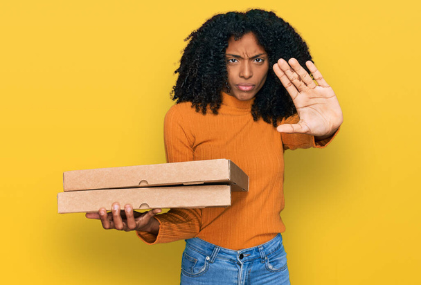Chica afroamericana joven sosteniendo caja de pizza de entrega con mano abierta haciendo stop sign con expresión seria y segura, gesto de defensa  - Foto, Imagen
