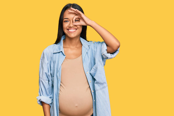 Bella donna ispanica in attesa di un bambino mostrando pancia incinta sorridente felice facendo ok segno con mano sugli occhi guardando attraverso le dita  - Foto, immagini
