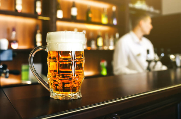Glass of beer with foam is standing on the wooden bar counter. Old pub with bartender cleaning the dishes on background. Leisure, fun and friends concepts photo. - Foto, Imagen