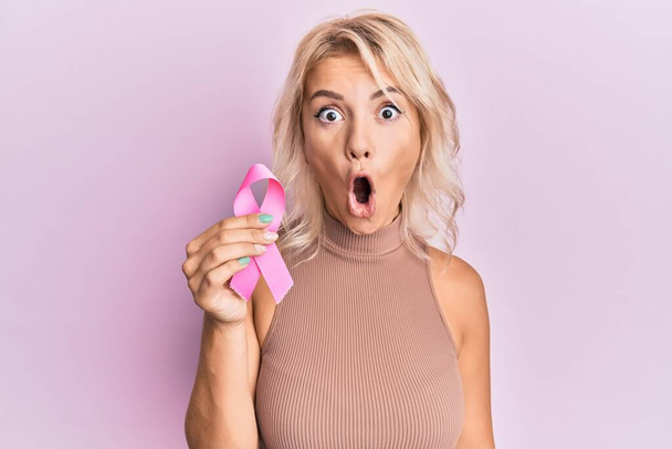 Young blonde girl holding pink cancer ribbon scared and amazed with open mouth for surprise, disbelief face  - 写真・画像