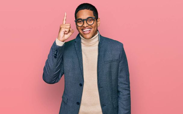 Junger gutaussehender hispanischer Mann mit Businessjacke und Brille, der mit erhobenem Zeigefinger auf eine erfolgreiche Idee hinweist. aufgeregt und glücklich. Nummer eins.  - Foto, Bild