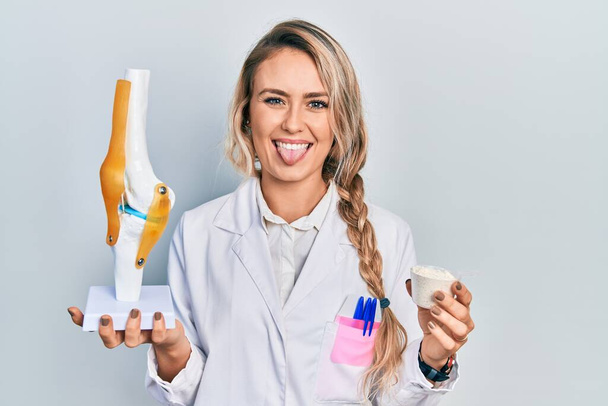 Bella giovane donna medico bionda in possesso di modello anatomico di articolazione del ginocchio e proteine in polvere attaccare lingua fuori felice con espressione divertente.  - Foto, immagini