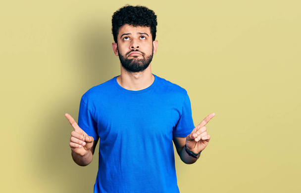 Young arab man with beard wearing casual blue t shirt pointing up looking sad and upset, indicating direction with fingers, unhappy and depressed.  - Fotografie, Obrázek