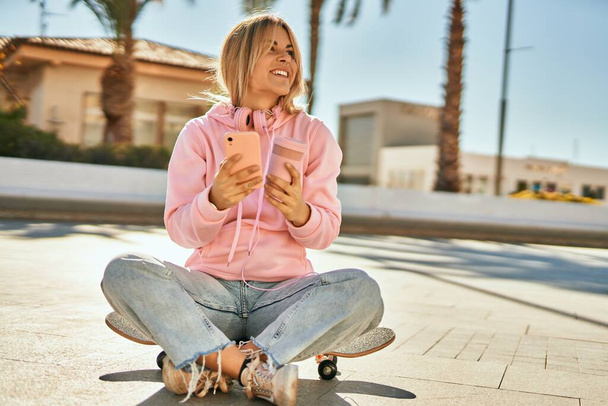 Jovem menina skatista loira usando smartphone e beber café sentado no skate - Foto, Imagem