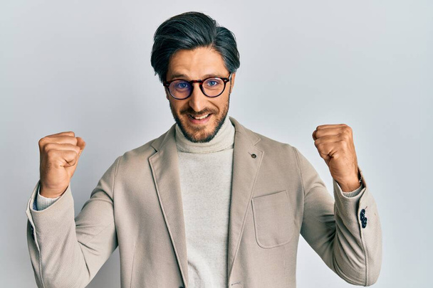 Fiatal spanyol férfi, aki üzleti kabátot és szemüveget visel, meglepetten és csodálkozva ünnepli a sikert felemelt karokkal és nyitott szemmel. győztes koncepció.  - Fotó, kép