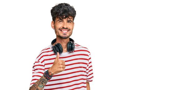 Молодой латиноамериканец слушает музыку, используя наушники, делая счастливый жест вверх рукой. одобрение выражения лица, смотрящего на камеру и показывающего успех.  - Фото, изображение