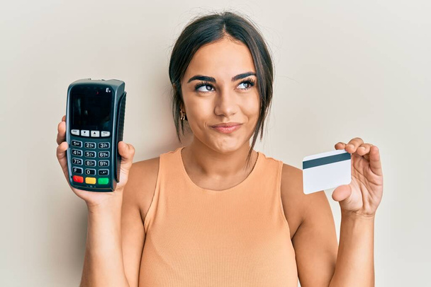 Mladá brunetka žena drží datafone a kreditní karty s úsměvem se dívá stranou a dívá se pryč myšlení.  - Fotografie, Obrázek