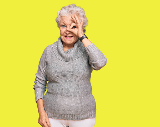 Femme âgée aux cheveux gris portant un pull d'hiver décontracté faisant un geste correct avec un sourire à la main, les yeux regardant à travers les doigts avec un visage heureux.  - Photo, image
