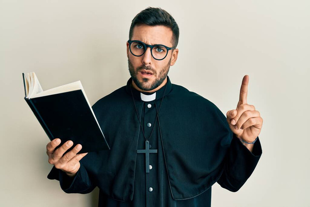 Junger hispanischer Priester, der mit erhobenem Finger die Bibel hält, ahnungslos und verwirrt. Zweifelhaftes Konzept.  - Foto, Bild