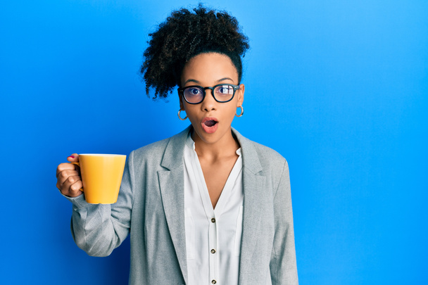 Junge afrikanisch-amerikanische Mädchen in Businesskleidung trinken eine Tasse Kaffee verängstigt und erstaunt mit offenem Mund für Überraschung, ungläubiges Gesicht  - Foto, Bild