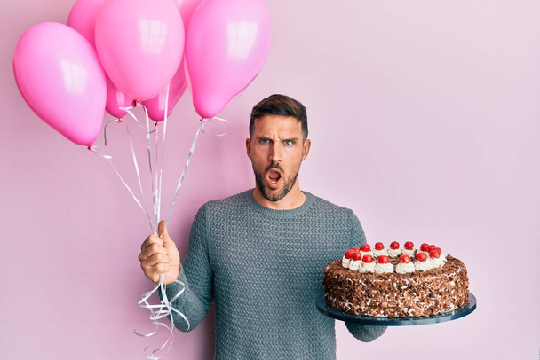 Homem bonito com barba comemorando aniversário com bolo segurando balões no rosto de choque, parecendo cético e sarcástico, surpreso com a boca aberta  - Foto, Imagem