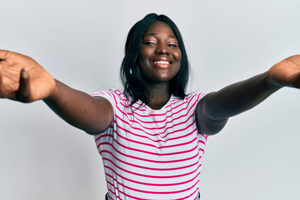 Africká mladá žena v neformálním pruhovaném tričku, dívá se do kamery a usmívá se s otevřenou náručí, aby ji objala. veselý výraz objímající štěstí.  - Fotografie, Obrázek