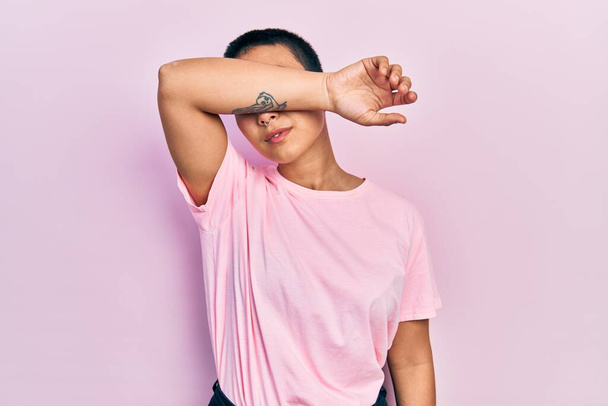Hermosa mujer hispana con el pelo corto vistiendo casual camiseta rosa cubriendo los ojos con el brazo, mirando serio y triste. concepto de ocultamiento y rechazo sin visión  - Foto, imagen
