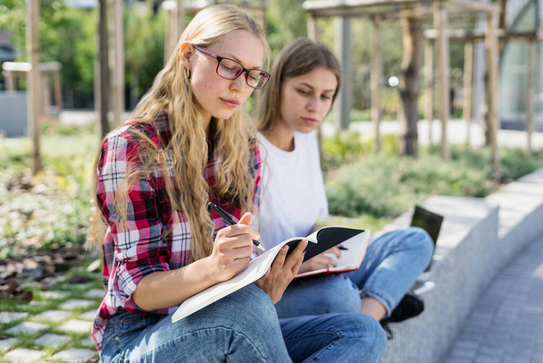 Education concept. Two smart and young university student girl study together, making scientific homework project, spending time near campus, writing notes in copybooks - Photo, Image