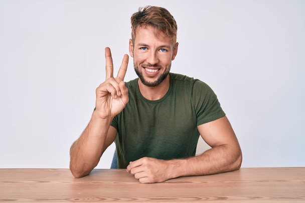 Young caucasian man wearing casual clothes sitting on the table showing and pointing up with fingers number two while smiling confident and happy.  - Photo, Image