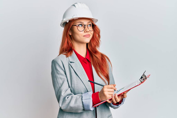 Young redhead woman wearing architect hardhat smiling looking to the side and staring away thinking.  - Foto, immagini