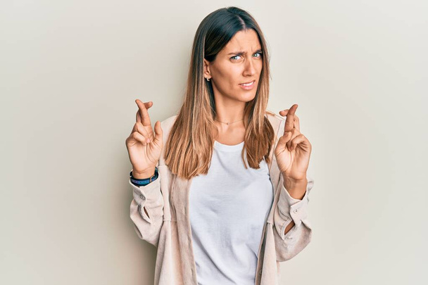 Brunette young woman doing fingers crossed gesture clueless and confused expression. doubt concept.  - Photo, Image