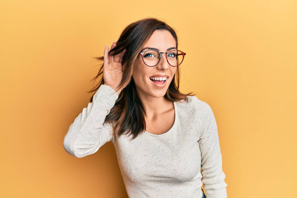 Jonge brunette vrouw draagt casual kleding en glazen glimlachen met hand over oor luisteren en horen van geruchten of roddels. doofheid.  - Foto, afbeelding