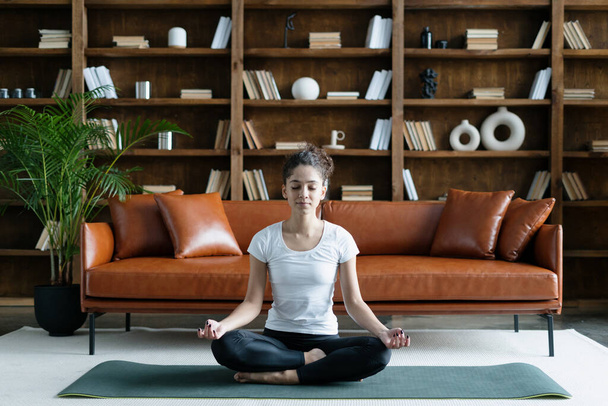 Nyugodt afro-amerikai nő visel sportruházat ül meditációs pózban, csinál jóga edzés sport szőnyeg, lélegzik mélyen edzés előtt. Fiatal felnőtt lány tölti nap otthon - Fotó, kép