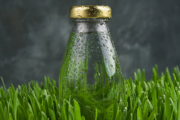 Bottle with yellow lid is covered with drops of moisture. Dew on glass in green grass. Natural freshness of morning, thirst quenching, motivation to drink - Photo, Image