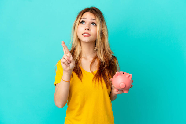 Teenager blonde girl holding a piggybank over isolated blue background with fingers crossing and wishing the best - Foto, afbeelding