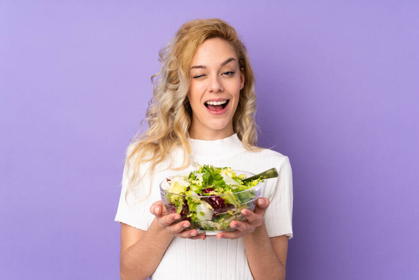Young blonde woman holding salad isolated on purple background - Фото, зображення