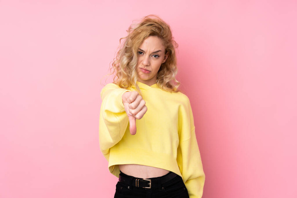 Молодая блондинка в толстовке на розовом фоне показывает большой палец вниз с отрицательным выражением лица - Фото, изображение