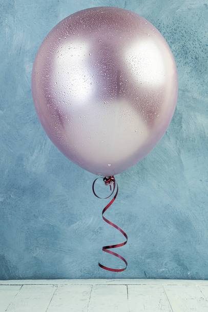 Pink balloon with tied red ribbon. It flies over wooden plank table against blue cement wall. Festive atmosphere of birthday party, wedding ceremony - Photo, Image