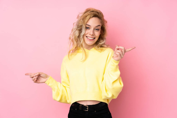 Junge blonde Frau trägt ein Sweatshirt isoliert auf rosa Hintergrund zeigt mit dem Finger auf die seitlichen und glücklich - Foto, Bild
