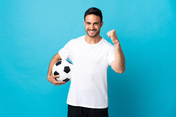 Jeune bel homme isolé sur fond bleu avec ballon de football célébrant une victoire - Photo, image