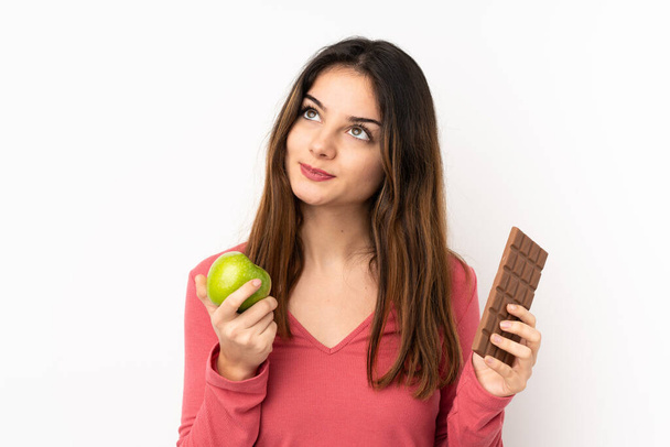 Молода біла жінка ізольована на рожевому тлі має сумніви, приймаючи шоколадну таблетку в одній руці і яблуко в іншій
 - Фото, зображення
