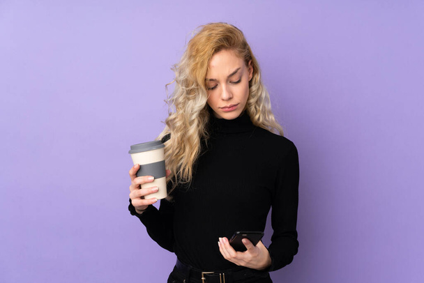 jonge blonde vrouw geïsoleerd op paarse achtergrond houden koffie mee te nemen en een mobiele - Foto, afbeelding
