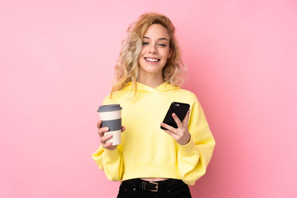 Jovem loira vestindo uma camisola isolada no fundo rosa segurando café para levar e um celular - Foto, Imagem