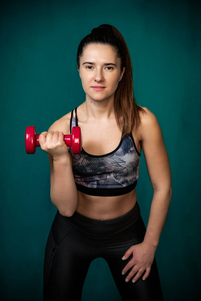 Sporty woman does the exercises with red dumbbells. Photo of young woman in sportswear on dark green background. Strength and motivation. - Fotó, kép