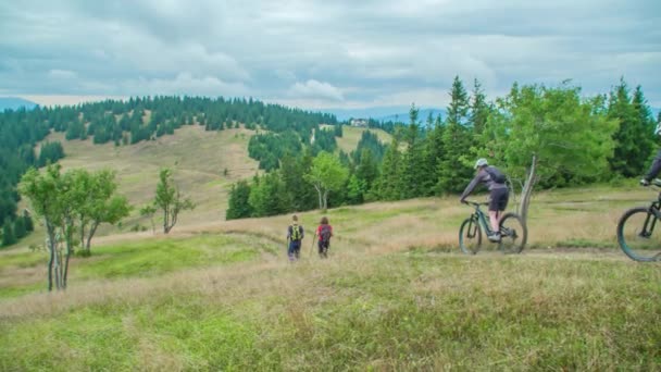 A couple of backpackers slowly go down the hill along the path, overtaking two cyclists. At backstage is a spruce forest with hills and a cloudy sky. - Footage, Video