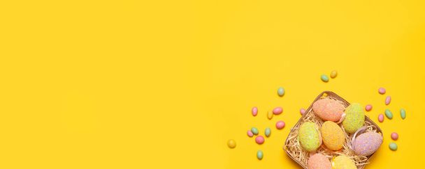 Festive basket with colored Easter eggs and tasty colored Candy jelly beans on a yellow background with copy space, top view, flat lay. - Photo, image
